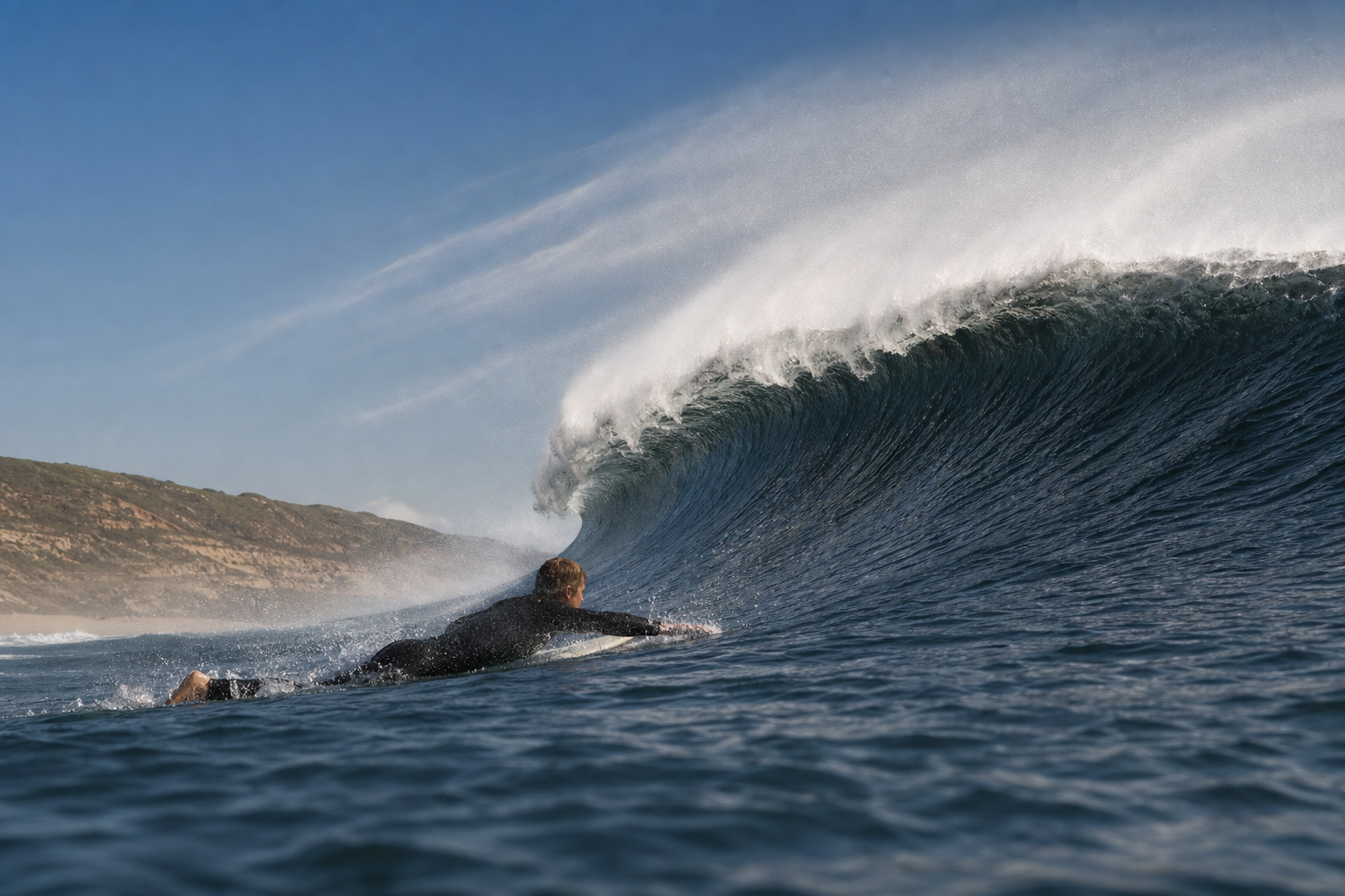 A prone surfer paddles toward a steep wave as heavy crest spray blows left toward the surfer and the distant shoreline.